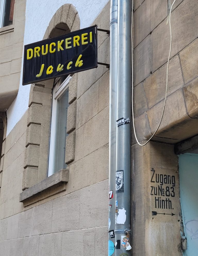 Schwarzes Schild mit der Aufschrift "Druckerei Jauch" an einer Gebäudeaußenwand mit Hinweis zu Hinterhaus.